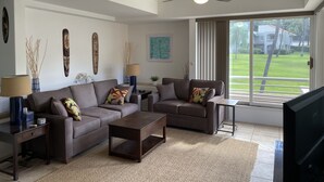 A modern living room with a sofa set, coffee table, and a TV. There is a ceiling fan and a large window with a view of a green landscape. - Beautiful - Palms at Wailea 1802 (Kihei)