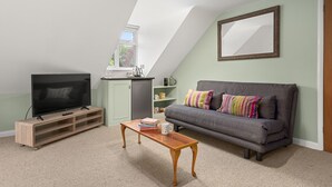 A living room with a television on a wooden stand, a grey sofa with colorful cushions, a wooden coffee table, and a small kitchen area with a window. - Chambers Place (Reading)