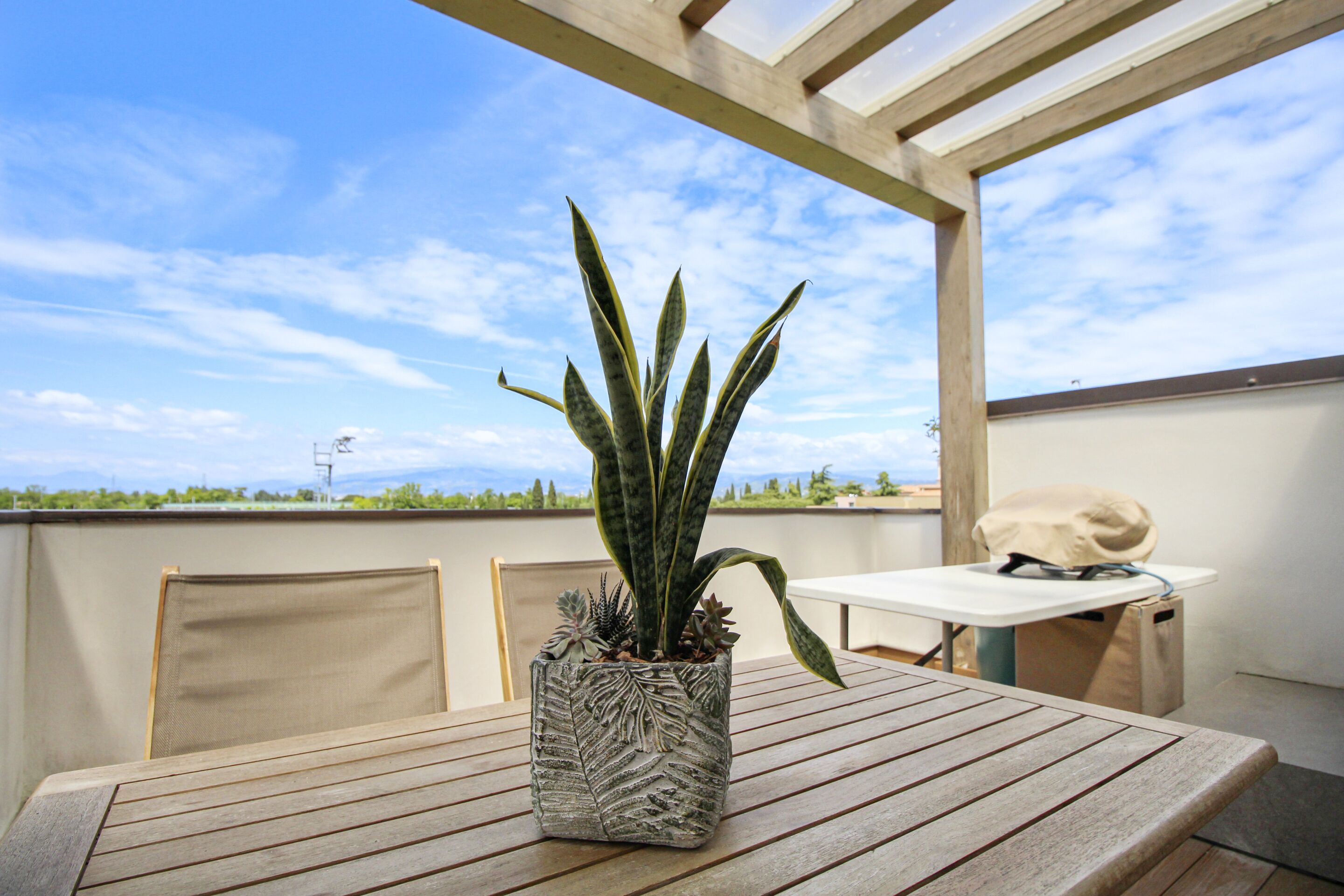 A wooden table with a potted plant, a chair, and a grill on a balcony with a clear sky.