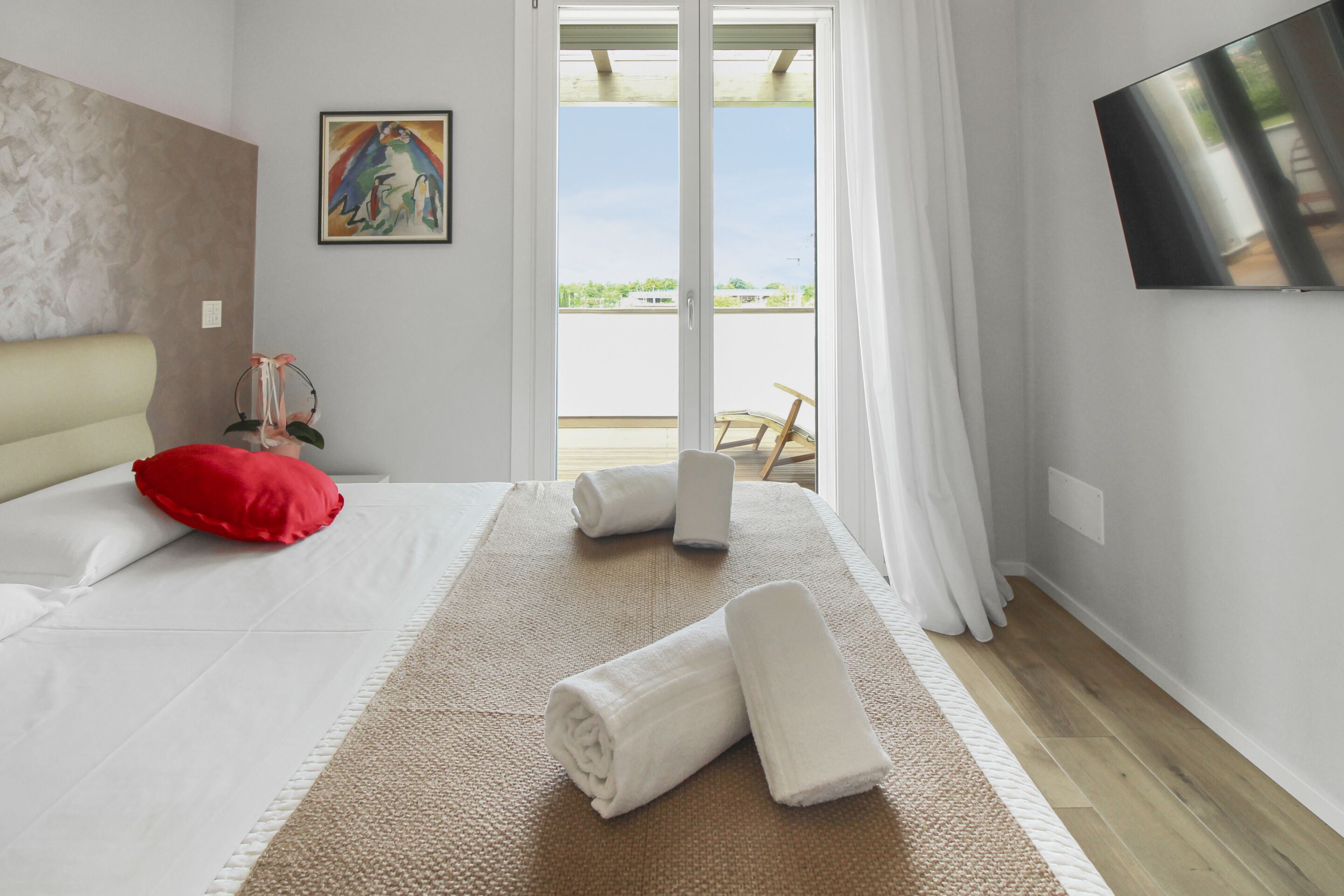 A hotel room with a bed, a red pillow, a painting on the wall, a TV mounted on the wall, and a view of the outdoors through a sliding door.
