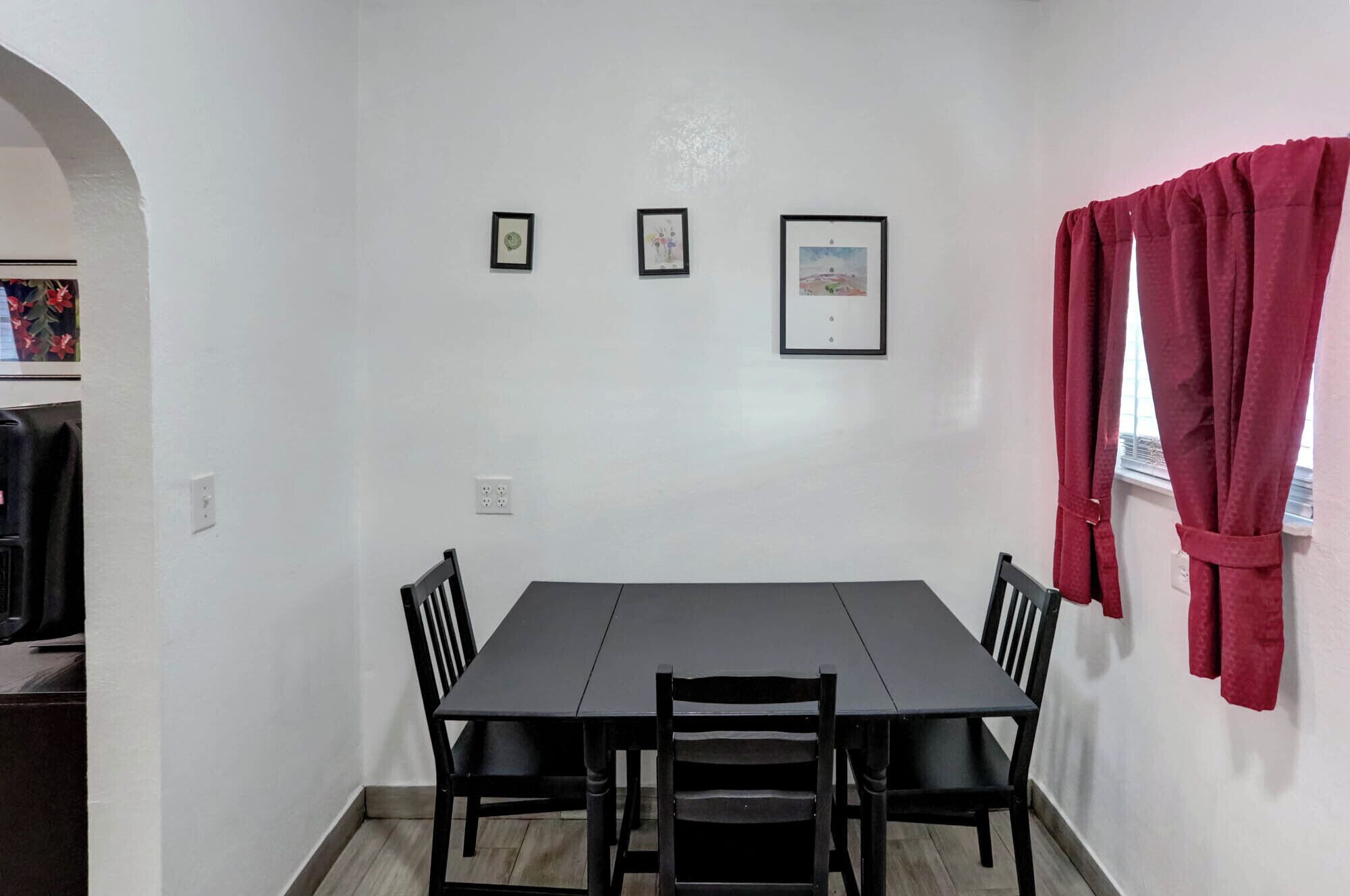 Un comedor con una mesa y cuatro sillas, una ventana con cortinas rojas y tres cuadros en la pared.