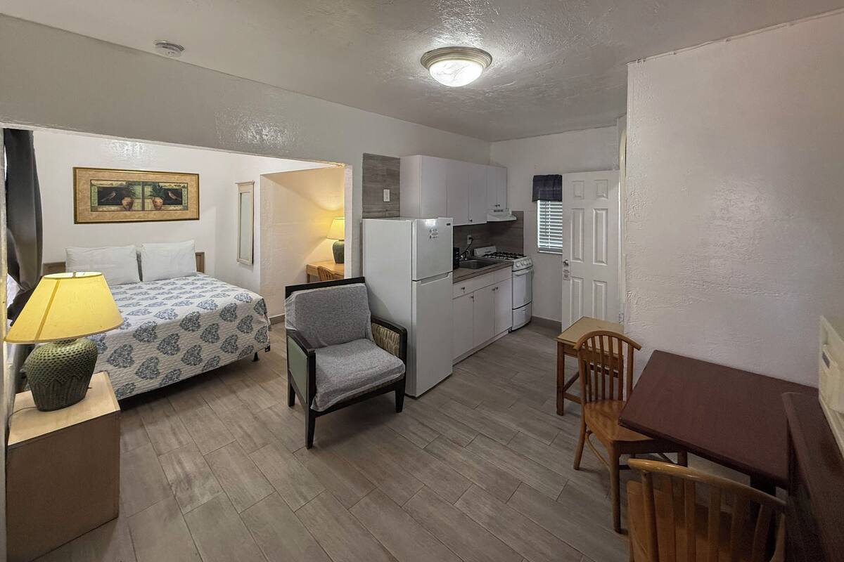 A compact room with a bed, a chair, a small table, a kitchenette, and a door.