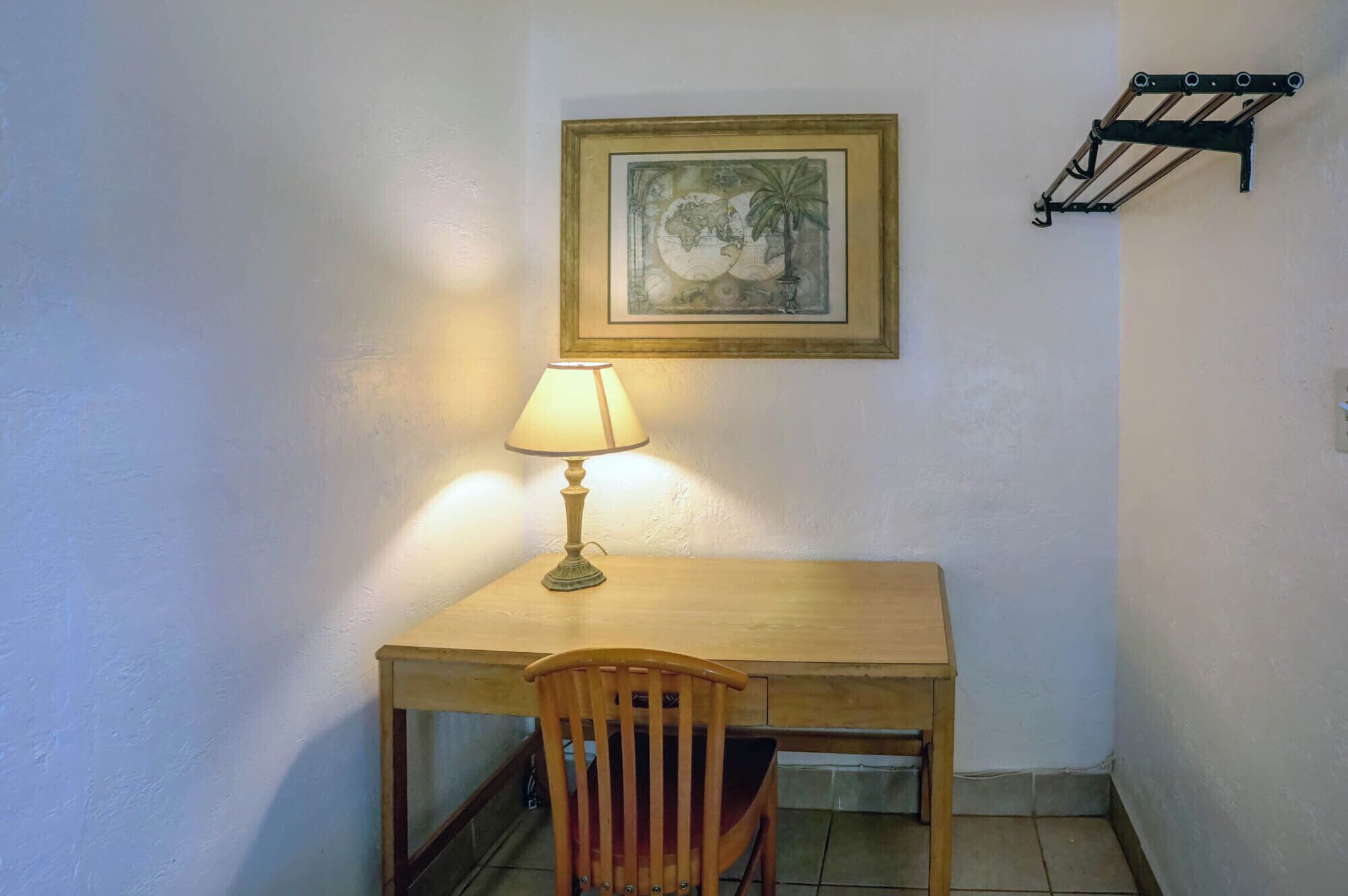A small, well-lit room with a wooden desk and chair, a lamp, and a framed picture on the wall.