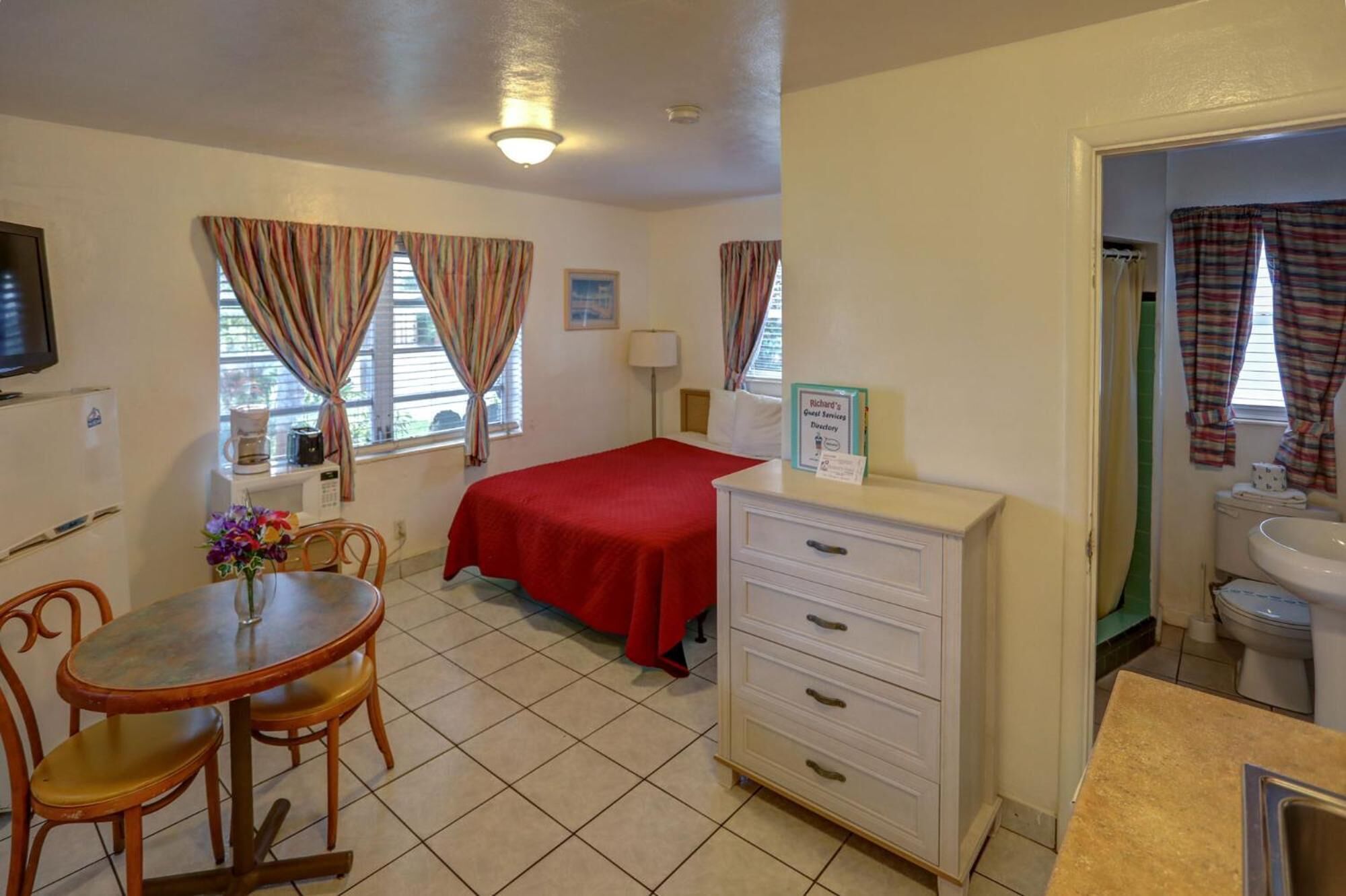 A small, clean room with a bed, a desk, a chair, a table with a vase of flowers, and a sign indicating guest services.