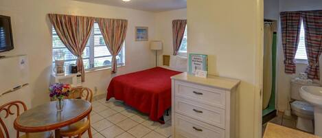 A small, clean room with a bed, a desk, a chair, a table with a vase of flowers, and a sign indicating guest services.