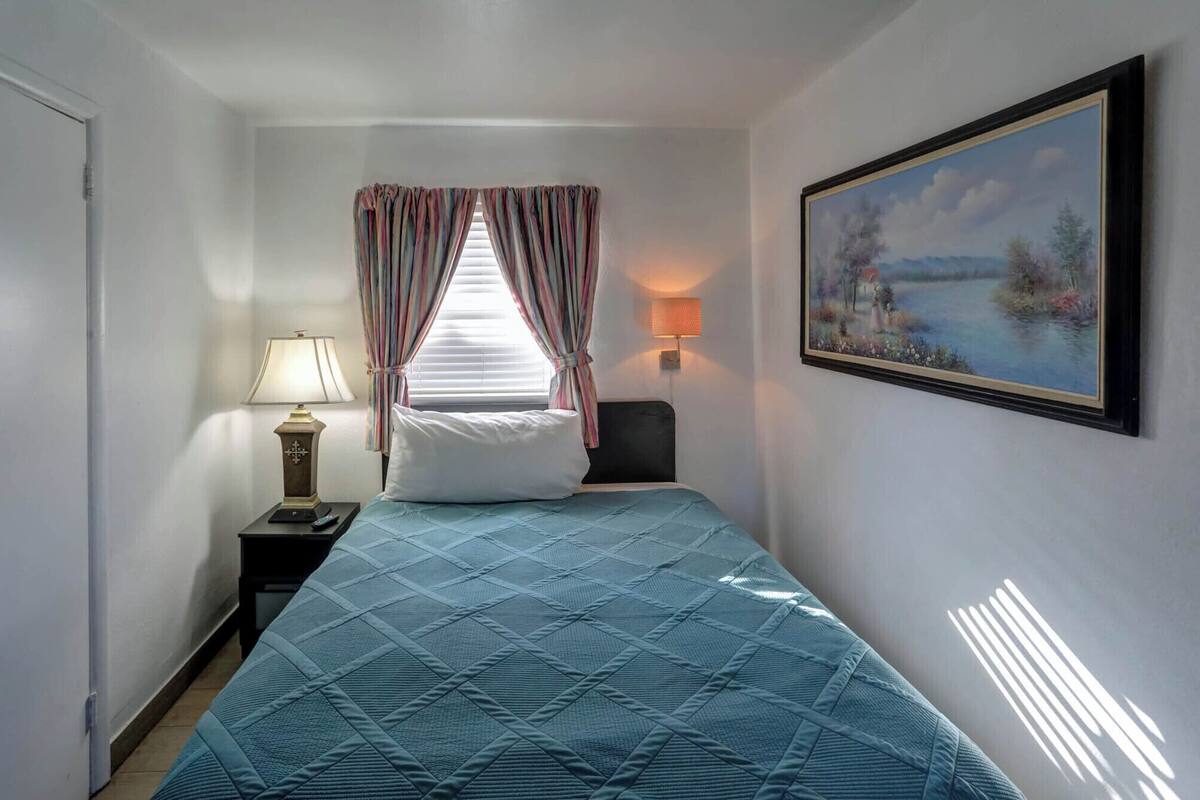 A hotel room with a bed, a nightstand, a lamp, a painting, and a window with curtains.