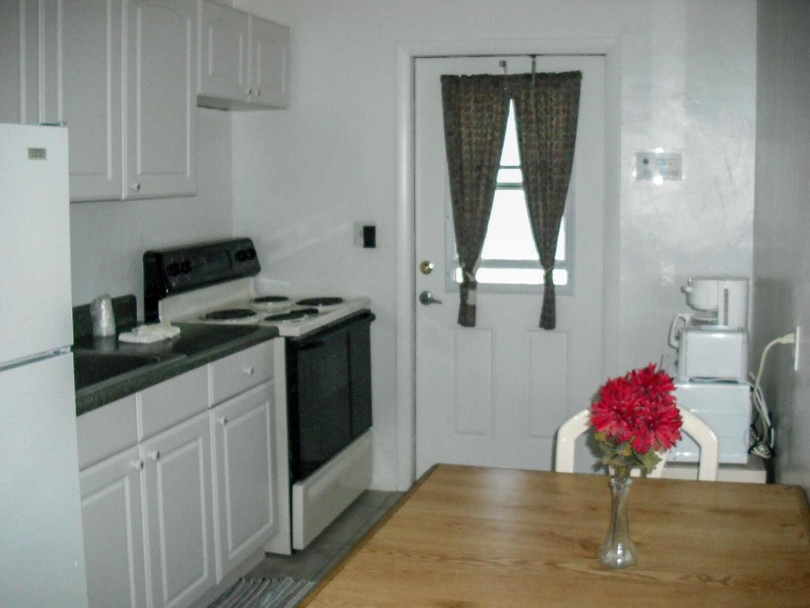 A small kitchen with white cabinets, a stove, a coffee maker, and a vase with red flowers on a wooden table.