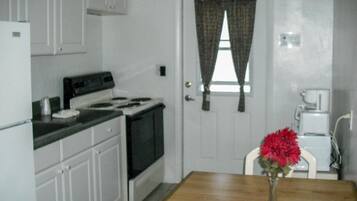 A small kitchen with white cabinets, a stove, a coffee maker, and a vase with red flowers on a wooden table.