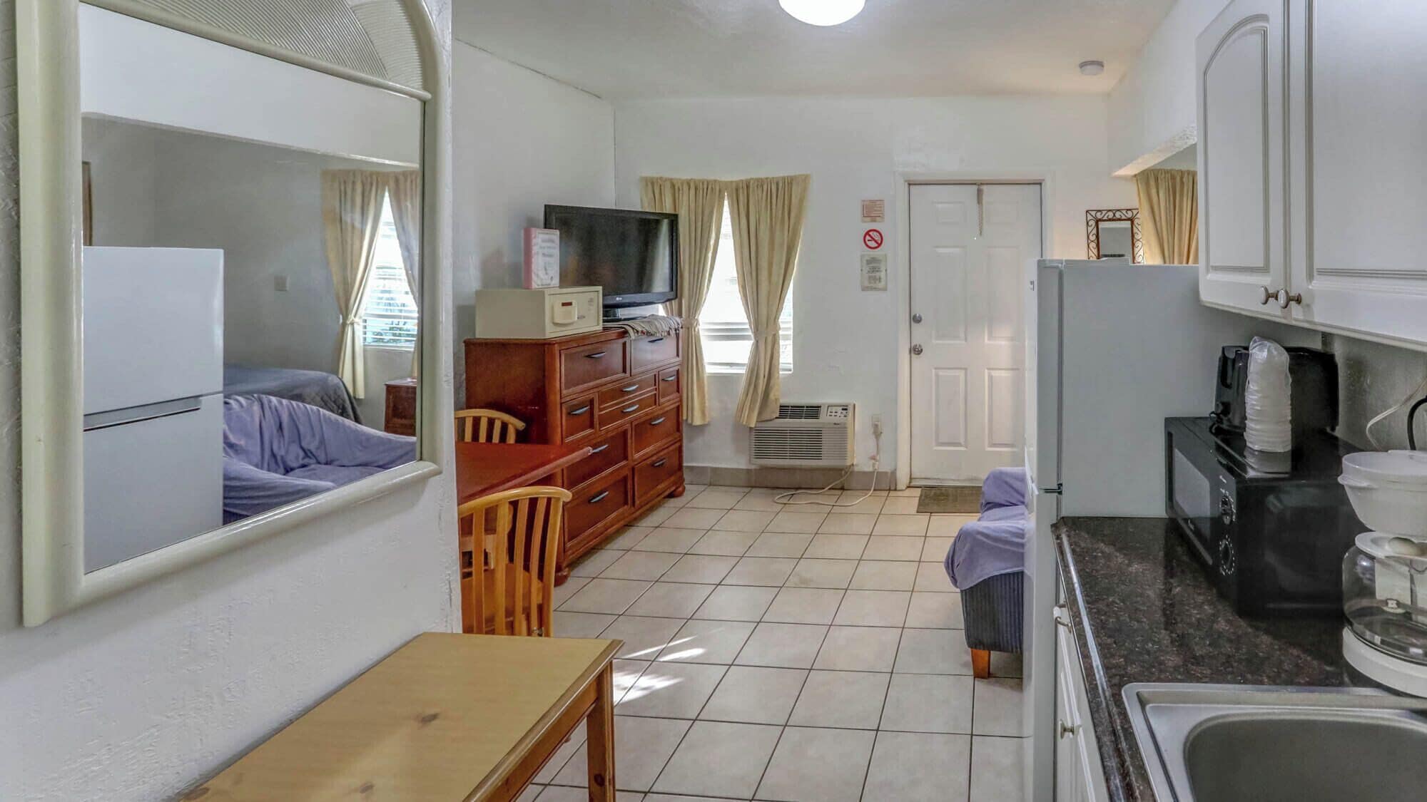 A compact room with a bed, a wooden dresser, a television, a kitchenette, and a door.