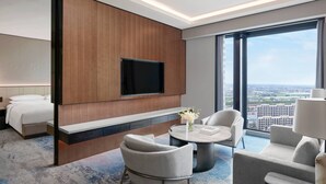 Een moderne hotelkamer met een bed, een flatscreen televisie bevestigd aan een houten paneel, een ronde salontafel met stoelen en een groot raam met uitzicht op de stad.