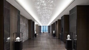 Een moderne hotelgang met een lange, rechte indeling, donker houten vloeren, glazen vitrines met witte vazen en een unieke plafondlamp.