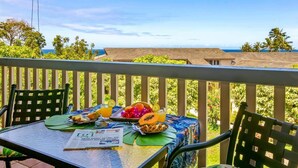 Ein Balkon mit einem Frühstückstisch, gedeckt mit Früchten und einer Zeitung, mit Blick auf eine schöne Landschaft mit Palmen und einem Gebäude.