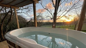 Un jacuzzi con cubierta de madera y barandilla metálica, rodeado de árboles y una vista al atardecer.