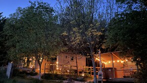 Una cabaña rústica de madera con luces colgantes, rodeada de árboles y un área con césped.