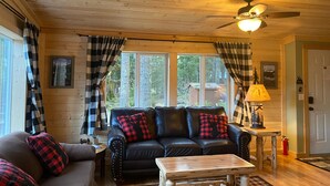 A cozy cabin interior with a leather sofa, plaid curtains, and a ceiling fan.