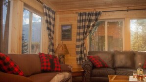 A cozy cabin interior with two sofas, a wooden coffee table, and large windows with plaid curtains.
