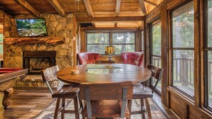 A cozy cabin interior with a stone fireplace, a round wooden dining table, and large windows offering a view of the forest. - Blue Ridge - Creek Front Paradise w/great outdoors (Blue Ridge)