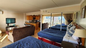 Habitación de hotel con dos camas, un sofá, televisión, kitchenette y balcón con vista a la ciudad.