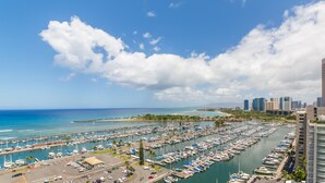 Un puerto deportivo con numerosos barcos amarrados, un cielo azul despejado y el horizonte de una ciudad al fondo.