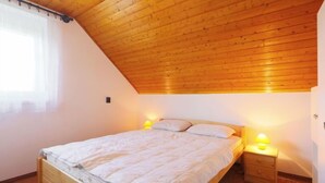 A bedroom with a sloped wooden ceiling, a wooden bed frame, a bedside table with a lamp, and a window with sheer curtains. - Vacation home Akazie by Interhome (Balatonmariafurdo)