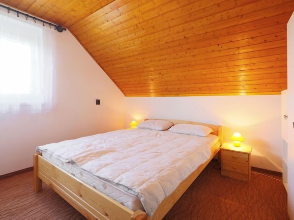 Et soveværelse med skråt træloft, en seng med træramme, et sengebord med lampe og et vindue med lette gardiner.
