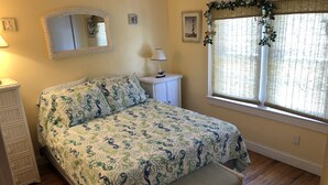 A bedroom with a bed, a nightstand, a wicker bench, a window with curtains, and a mirror on the wall. - Beach House in Ocean Beach, Fire Island (Ocean Beach)