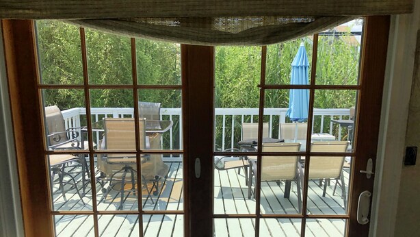 A view through a glass door to an outdoor deck with a white railing, several chairs, and a blue umbrella. - Beach House in Ocean Beach, Fire Island (Ocean Beach)