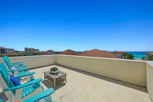 A balcony with turquoise chairs and a small table, overlooking a coastal area with buildings and the sea. - Beaches Be Sippin' - NEW! Ocean Views with Private Pool & Rooftop Deck (Destin)