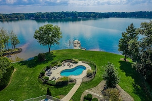 A swimming pool surrounded by a well-maintained lawn, with a dock extending into a lake.