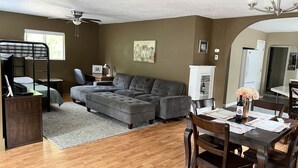 A modern living room with a sectional sofa, a dining table with chairs, a bunk bed, a ceiling fan, and a kitchen area visible through an archway. - Entire Home & Fenced Yard For Family & Pets (Independence)