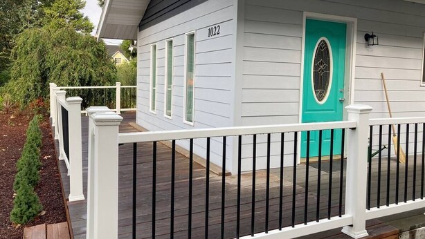 A small, single-story building with a white exterior and a turquoise door. It has a white porch with a black railing and a small garden area with a wooden planter. - Entire Home & Fenced Yard For Family & Pets (Independence)
