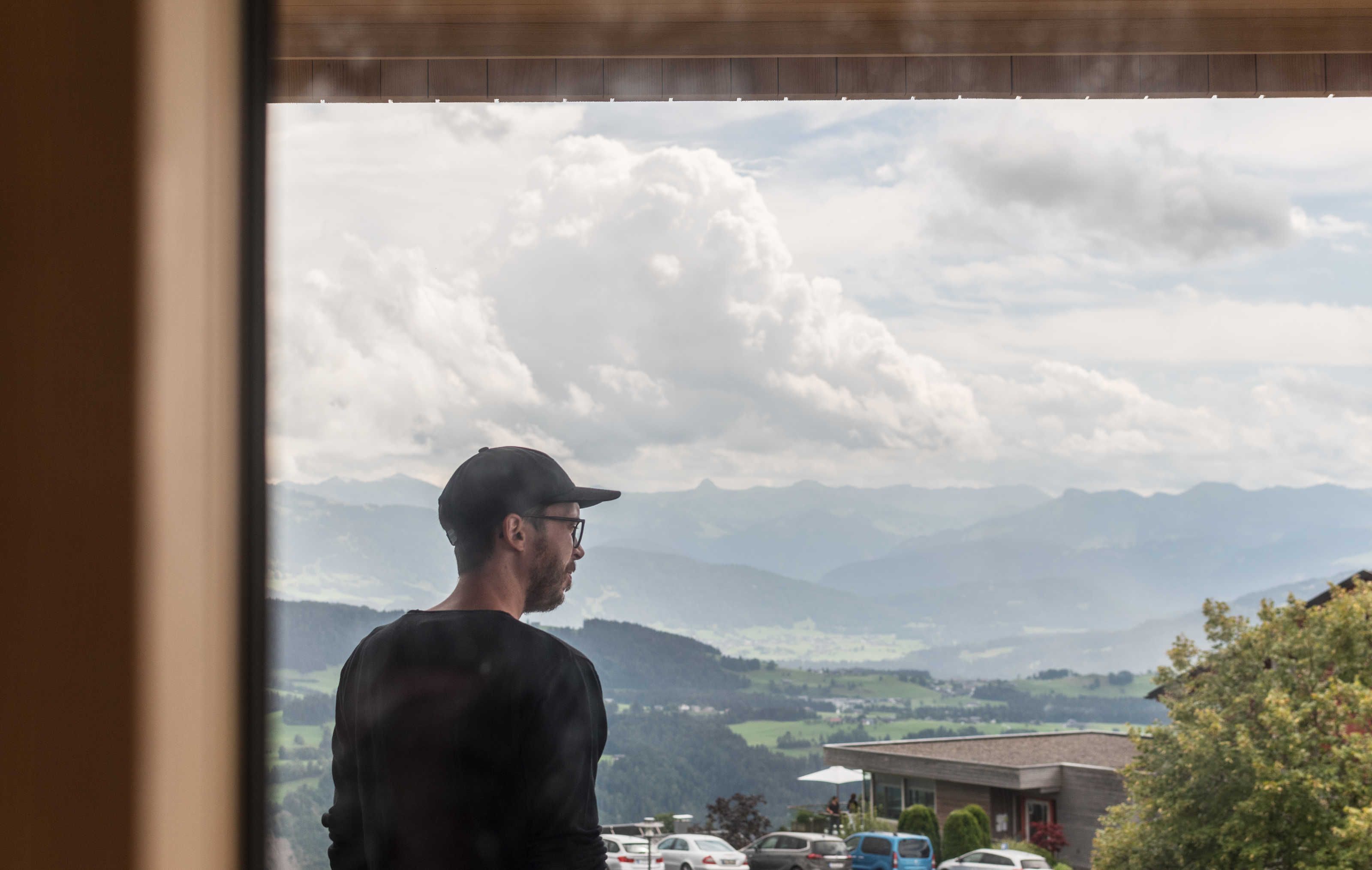 A person standing indoors, looking out at a mountainous landscape with clouds.