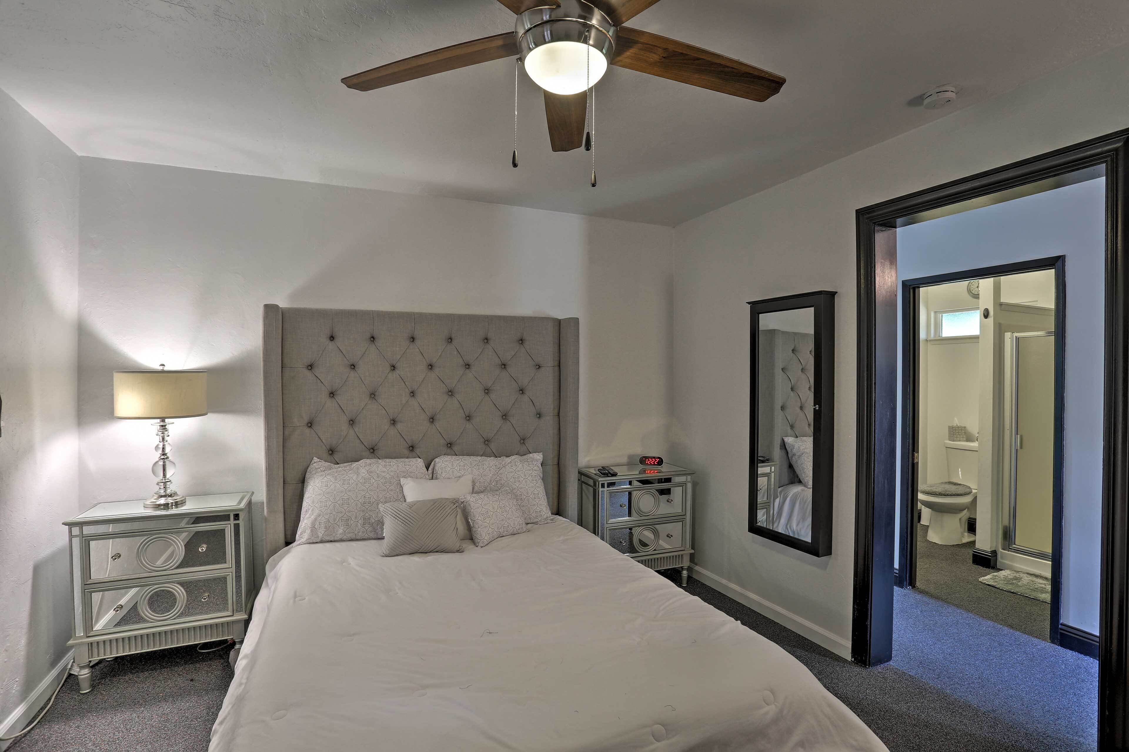 Een slaapkamer met een gestoffeerd hoofdeinde, een bed met wit beddengoed, een nachtkastje met een lamp, een nachttafeltje met een klok en een spiegel aan de muur.