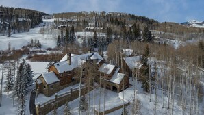 A large log cabin with multiple rooms, surrounded by snow-covered trees and a ski slope in the background. - GOLD HILL- Spacious, Luxurious Mountain Village Home (Mountain Village)
