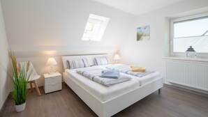 A modern bedroom with a large bed, bedside tables with lamps, a potted plant, and a window with a view of the roof.