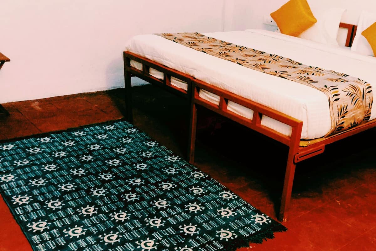 A bed with a patterned bedspread, two yellow pillows, and a wooden frame. There is a small table with a white object on it and a blue patterned rug on the floor.