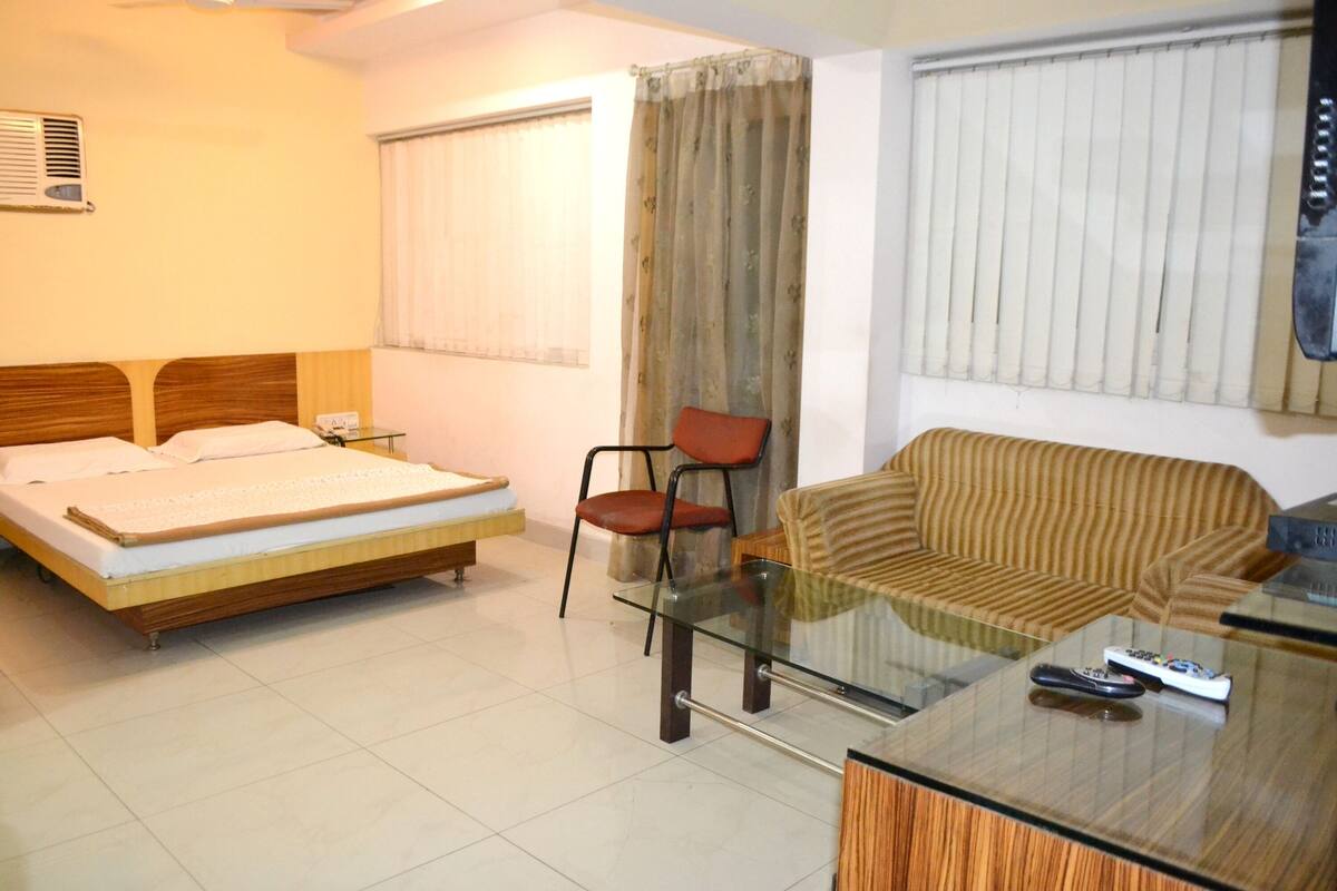 A hotel room with a bed, a glass table with remote controls, a sofa, and a chair.