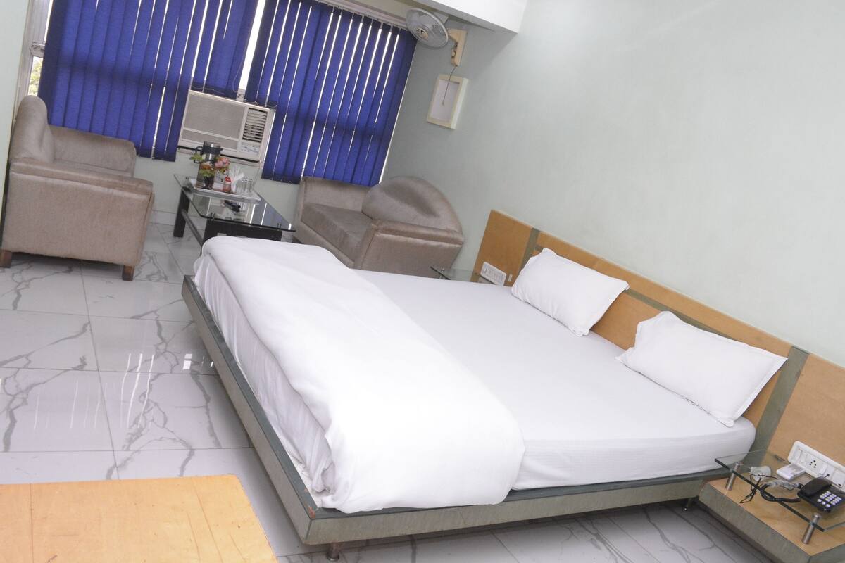 A hotel room with a bed, two chairs, a small table with a vase, and a wall-mounted air conditioner.
