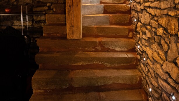 Uma escadaria de pedra leva a uma porta de madeira, com uma parede de pedra de um lado e uma estrutura de vigas de madeira do outro.