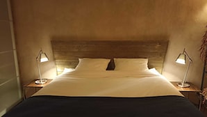 Cama bem arrumada com cabeceira de madeira, duas luminárias laterais e um cobertor escuro.