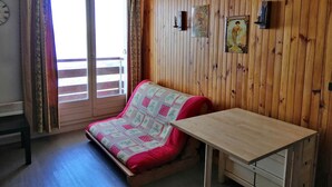 Een gezellige kamer met een houten lambrisering, een bed met een bedovertrek met dessin, een tafeltje en een raam met gordijnen.
