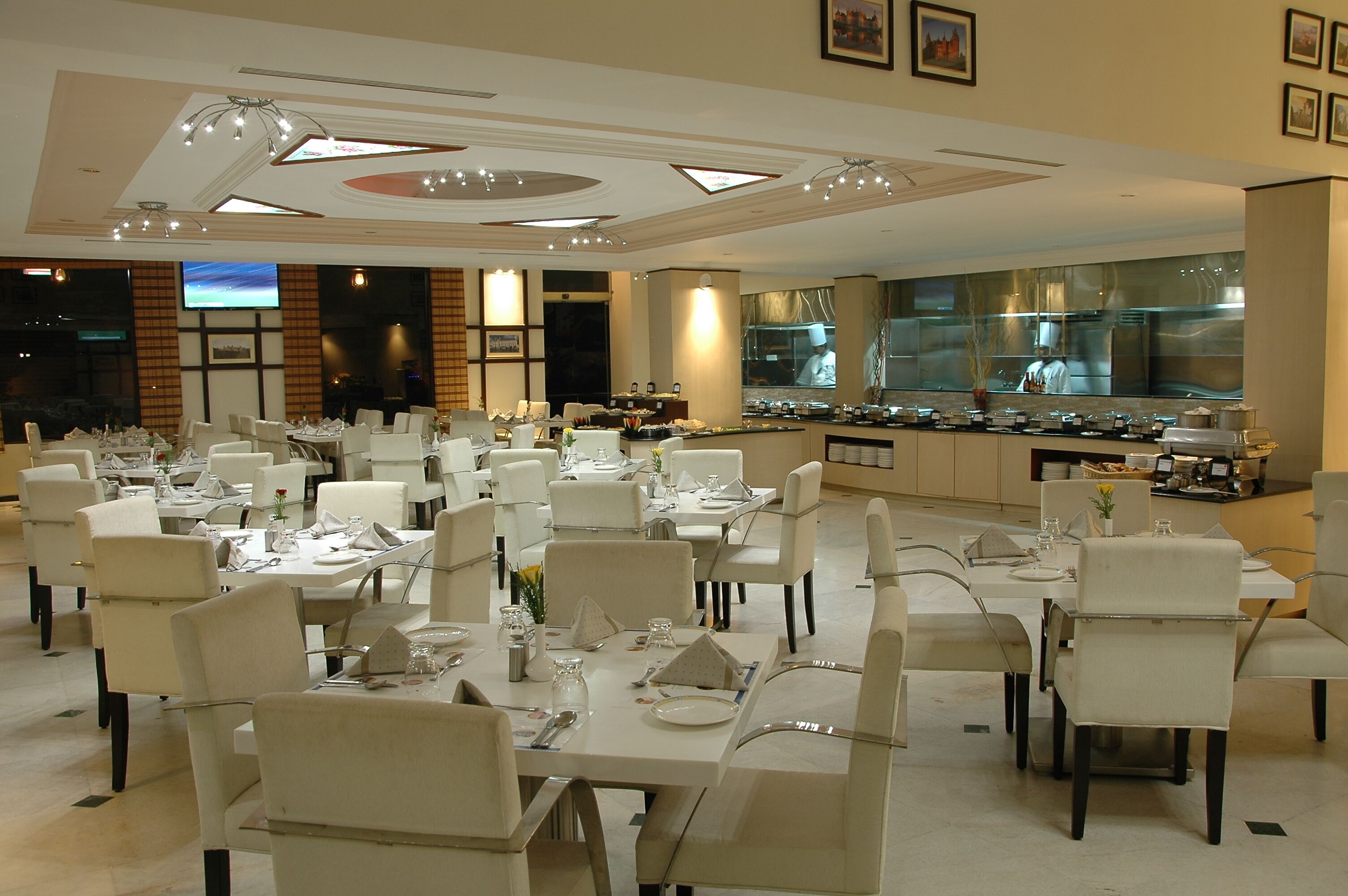 A spacious dining area with multiple tables set for guests, a large kitchen visible through the glass wall, and framed artwork on the walls.