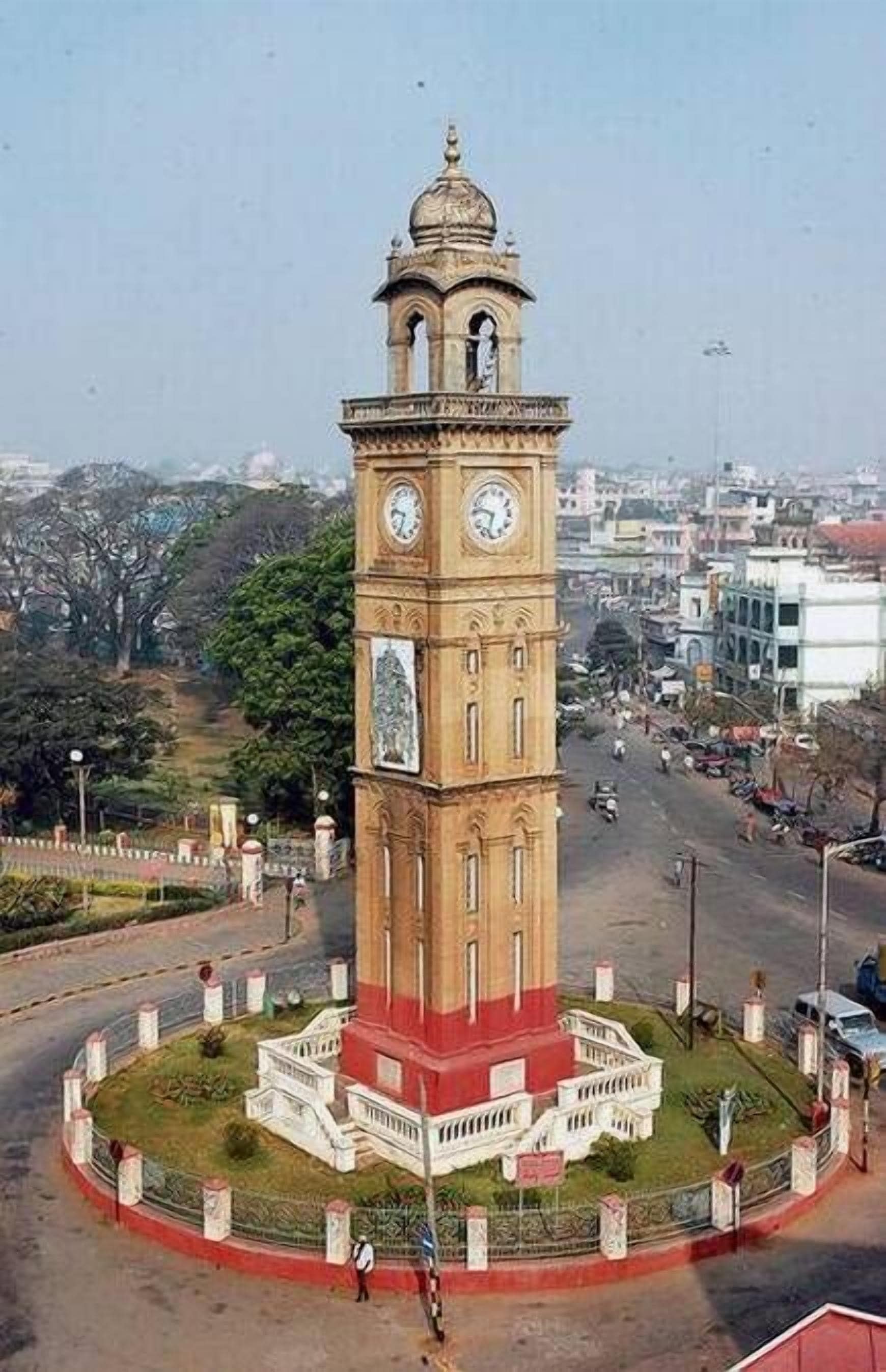 Une tour horloge avec une horloge centrale et une statue à sa base, entourée d’une place circulaire délimitée par une clôture.