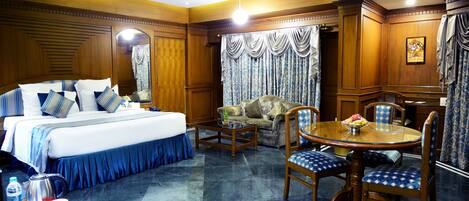 A hotel room with a large bed, a dining table with chairs, a coffee table, and a ceiling fan.