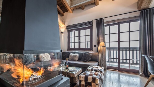 Una sala de estar acogedora con chimenea, piso de madera, un sofá con cojines y un amplio ventanal con cortinas.