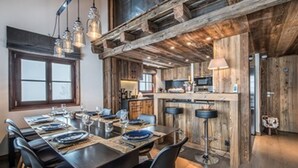 Un comedor rústico con una larga mesa dispuesta para seis comensales, vigas de madera en el techo y una zona de cocina con microondas y estufa.
