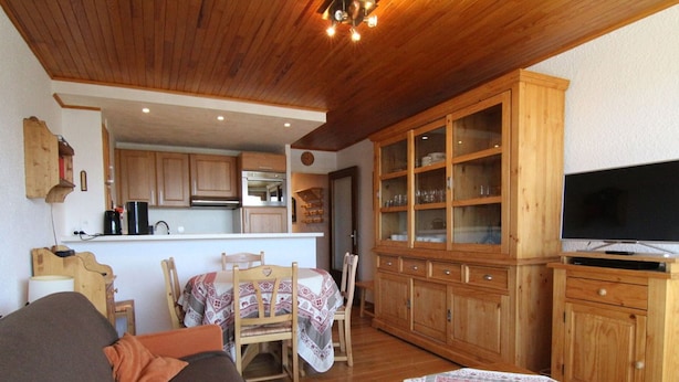Een gezellige woonkamer met een bank, een eettafel met stoelen, een houten kast met glazen deuren en een televisie op een standaard.