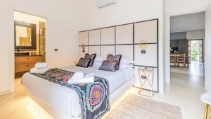 Een moderne slaapkamer met een groot bed, een nachtkastje, een spiegel en uitzicht op de woonkamer.