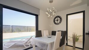 A modern dining area with a table and chairs, a clock on the wall, and a view of a swimming pool. - Villa Barzinu (Isla de Fraile) (Aguilas)