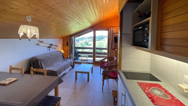 A cozy cabin interior with a wooden ceiling, a dining area with a table and chairs, a sofa, a kitchen area with a microwave, and a large window offering a view of the outdoors. - Studio cabin 5 pers. with south balcony and GARAGE, Les Saisies (Les Saisies)